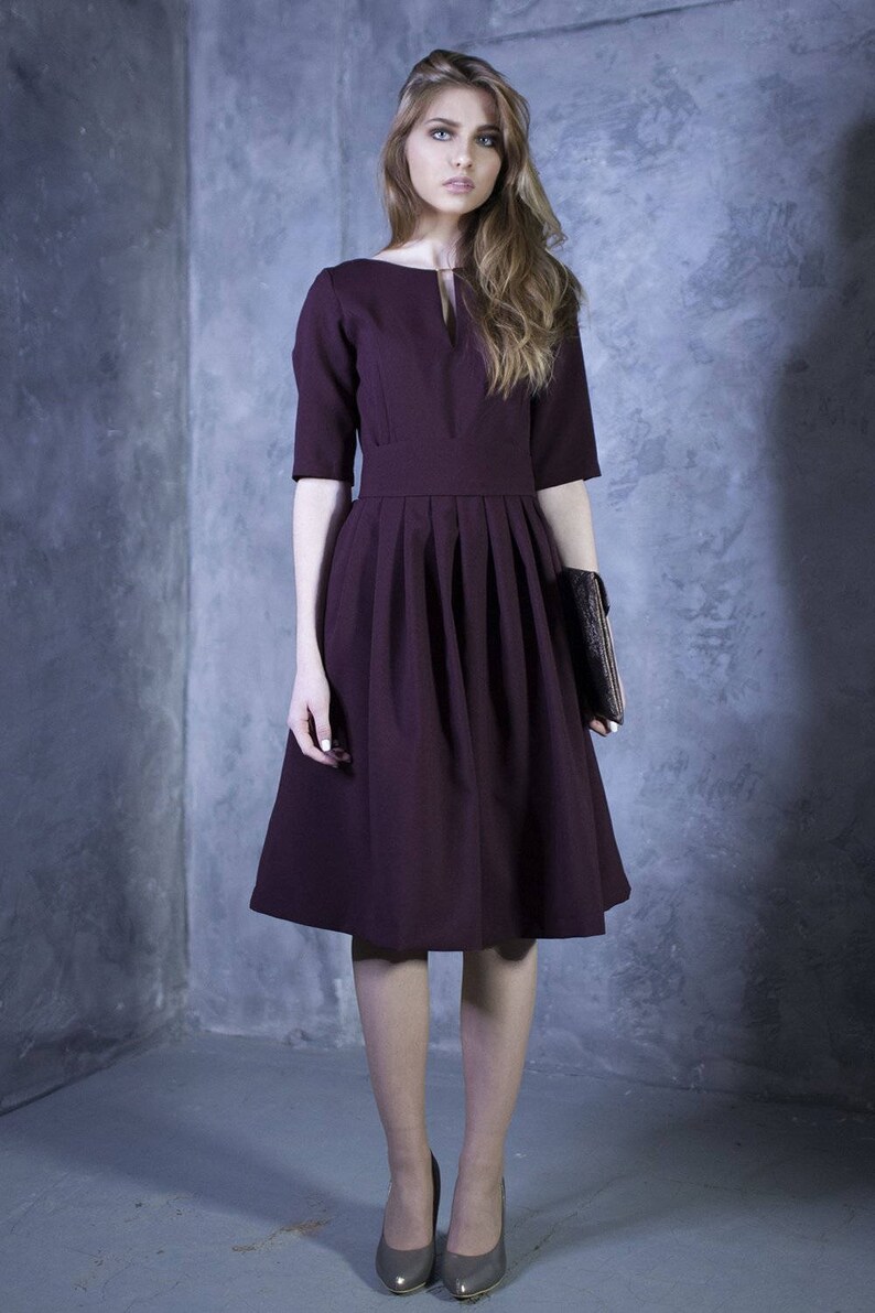 May include: A woman wearing a burgundy dress with a pleated skirt and a small clutch. She is standing in front of a gray wall.