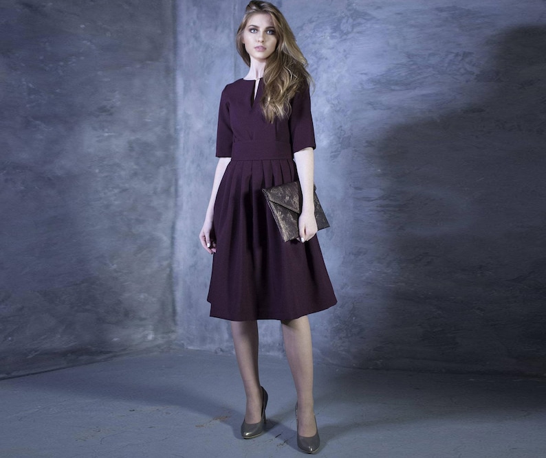 May include: A woman wearing a maroon dress with a V-neck and a pleated skirt. She is holding a brown clutch purse and wearing high heels.
