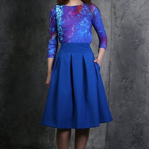 Pu&ograve; includere: Una donna indossa una camicia a maniche lunghe con motivo blu e viola e una gonna plissettata blu reale.