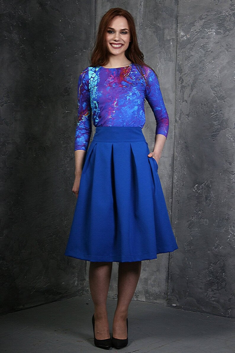 Pu&ograve; includere: Una donna indossa una camicia a maniche lunghe con motivo blu e viola e una gonna plissettata blu reale.