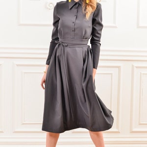 Pu&ograve; includere: Un abito midi grigio con un corpetto a camicia con colletto e una gonna lunga. L'abito ha una cintura a fiocco in vita e maniche lunghe. La modella indossa tacchi gialli.
