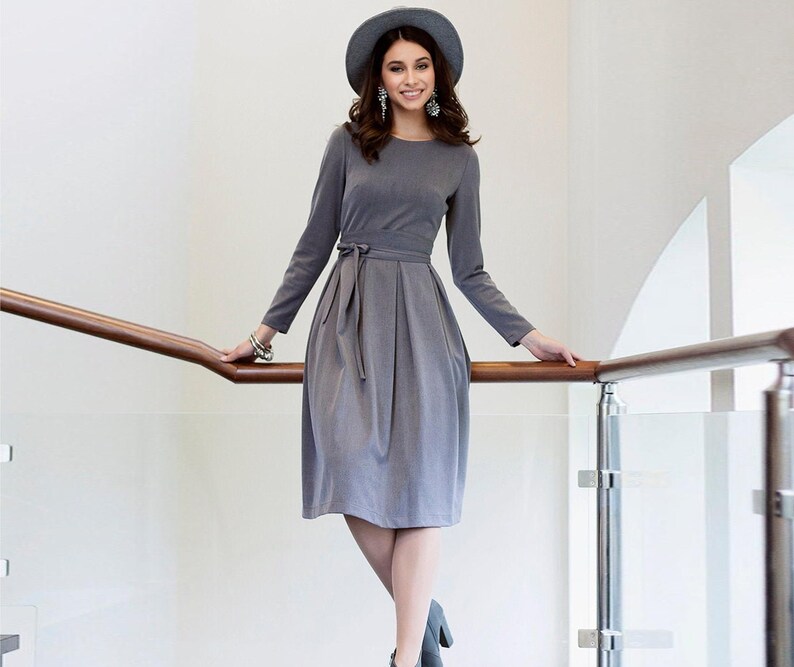 Puede incluir: Una mujer con un vestido gris con cintur&oacute;n de lazo y un sombrero gris. Est&aacute; de pie en una escalera con una barandilla de madera.