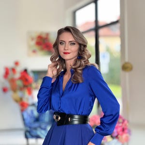 May include: A woman wearing a royal blue satin blouse with a black belt and a matching skirt. She has long, wavy blonde hair and is wearing blue earrings.