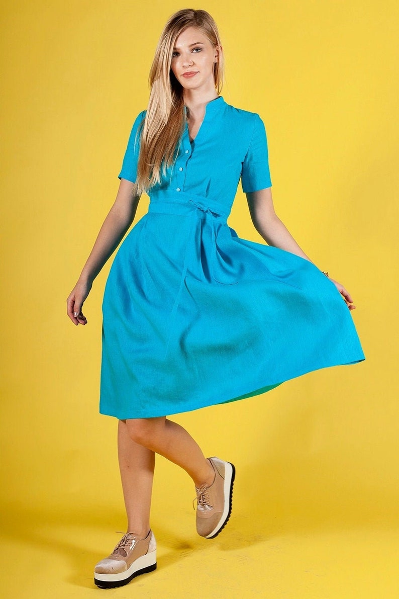 May include A woman wearing a turquoise blue dress with a white button-up collar and a matching belt. She is standing on a yellow background and wearing beige platform shoes.