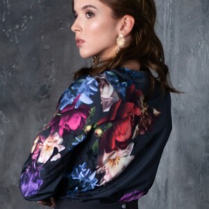 May include: A woman wearing a black blouse with a floral print and a black skirt. The blouse has a large floral print with pink, red, blue, and white flowers. The skirt is a simple black skirt with a pleated design.