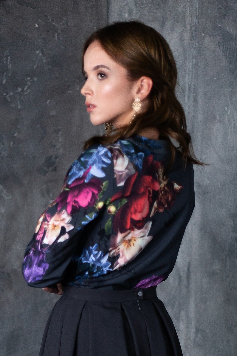 May include: A woman wearing a black blouse with a floral print and a black skirt. The blouse has a large floral print with pink, red, blue, and white flowers. The skirt is a simple black skirt with a pleated design.