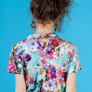 May include: A woman wearing a short-sleeved, light blue blouse with a floral print. The blouse has a collar and a keyhole opening in the back. She is also wearing a turquoise skirt.