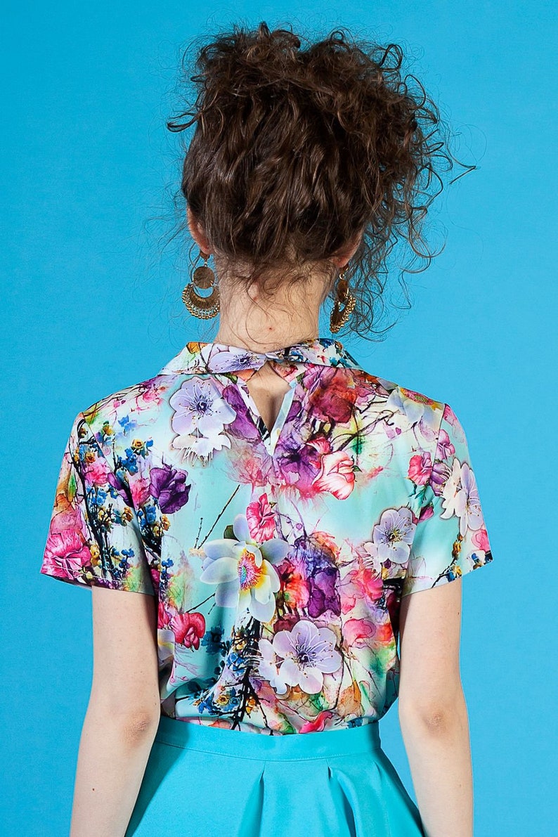 May include: A woman wearing a short-sleeved, light blue blouse with a floral print. The blouse has a collar and a keyhole opening in the back. She is also wearing a turquoise skirt.