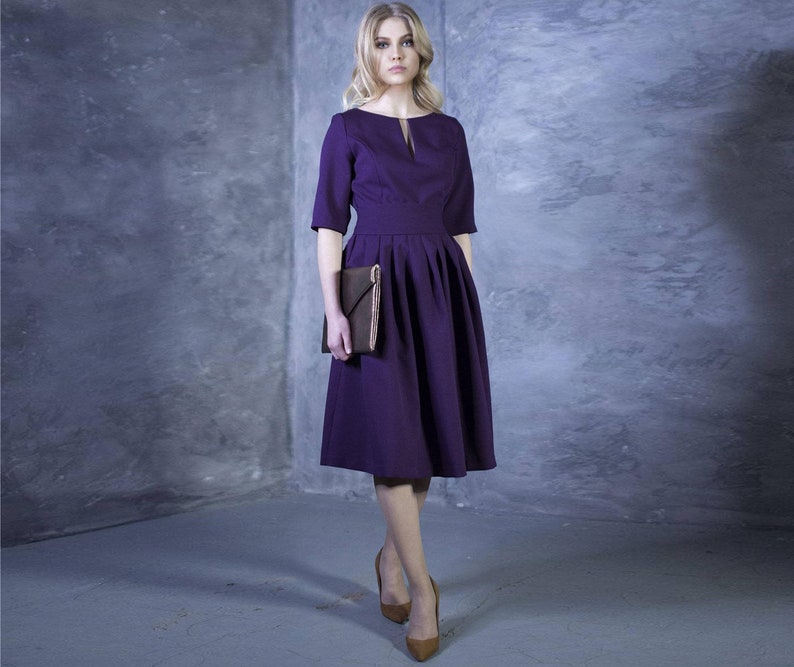 May include: A woman wearing a purple knee-length dress with a small V-neck and a pleated skirt. She is holding a brown clutch and wearing brown heels.