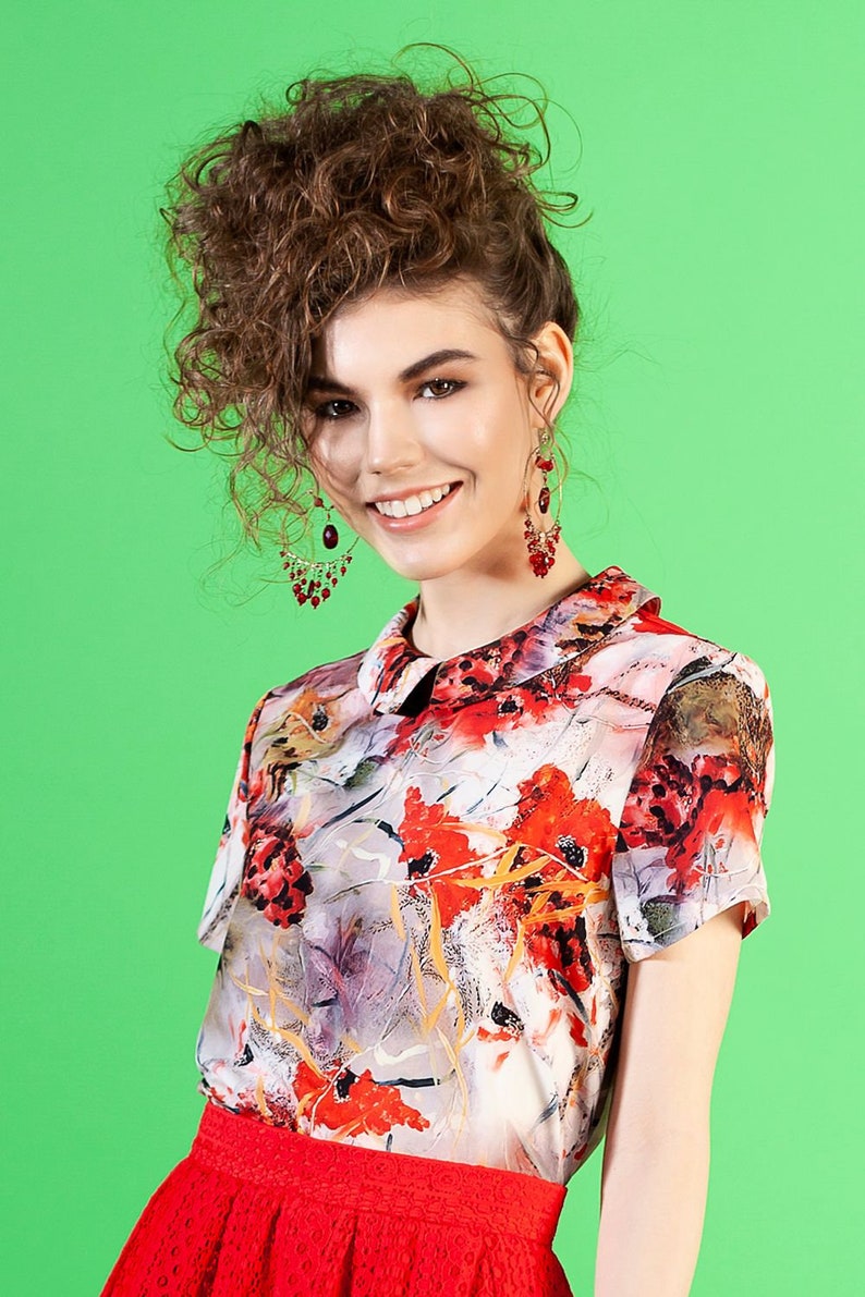 May include: A woman with curly brown hair wearing a white blouse with a floral print and a red skirt. She is wearing red earrings.