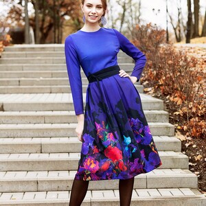 K&ouml;nnte beinhalten: Eine Frau in einem blauen Kleid mit einem Rock mit Blumenmuster und schwarzen Strumpfhosen. Sie steht auf einer Treppe vor einem Hintergrund aus Herbstlaub.