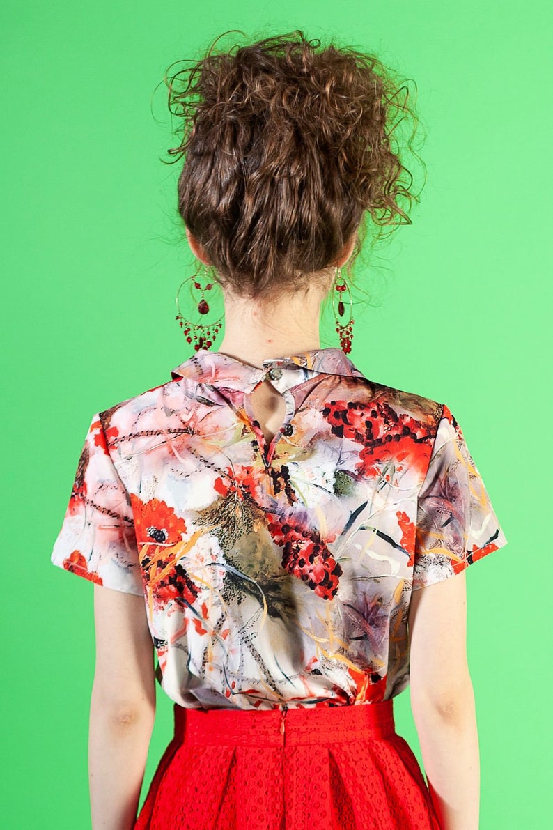 May include: A woman wearing a red skirt and a white blouse with a floral print. The blouse has a collar and short sleeves. The woman has curly brown hair and is wearing dangling earrings.