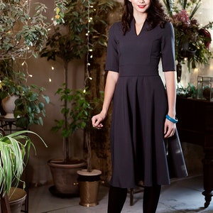 May include A woman wearing a dark gray dress with a v-neck and a flared skirt. She is wearing black tights and ankle boots. She is standing in a room with plants and string lights.