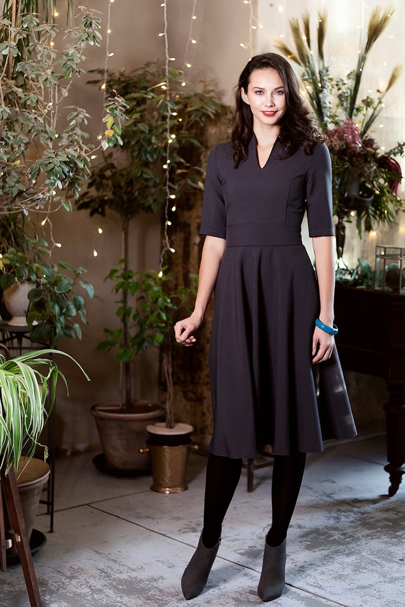 May include A woman wearing a dark gray dress with a v-neck and a flared skirt. She is wearing black tights and ankle boots. She is standing in a room with plants and string lights.
