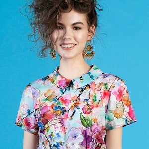 May include: A woman with curly brown hair is wearing a short-sleeved, light blue blouse with a floral print. The blouse has a white collar and a light blue skirt. She is wearing gold earrings.