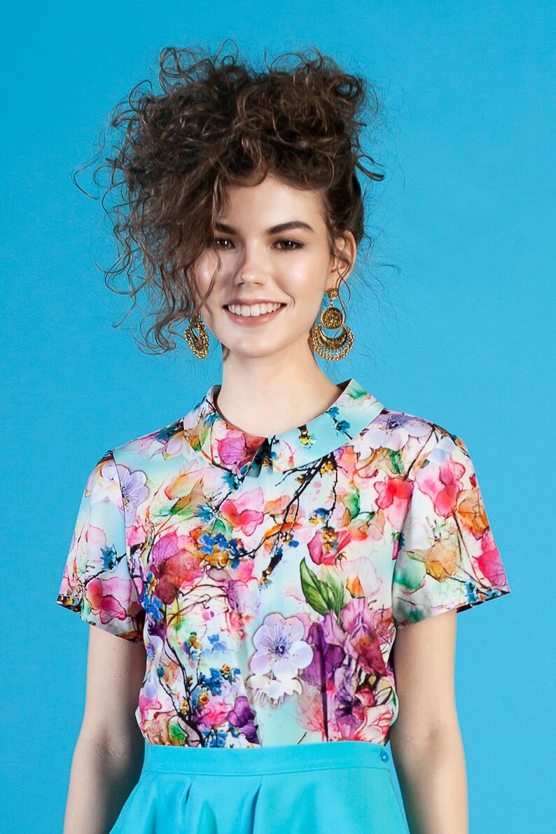 May include: A woman with curly brown hair is wearing a short-sleeved, light blue blouse with a floral print. The blouse has a white collar and a light blue skirt. She is wearing gold earrings.