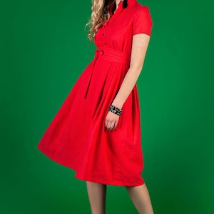 May include A woman wearing a red dress with a belt and short sleeves. She is standing on a green background.