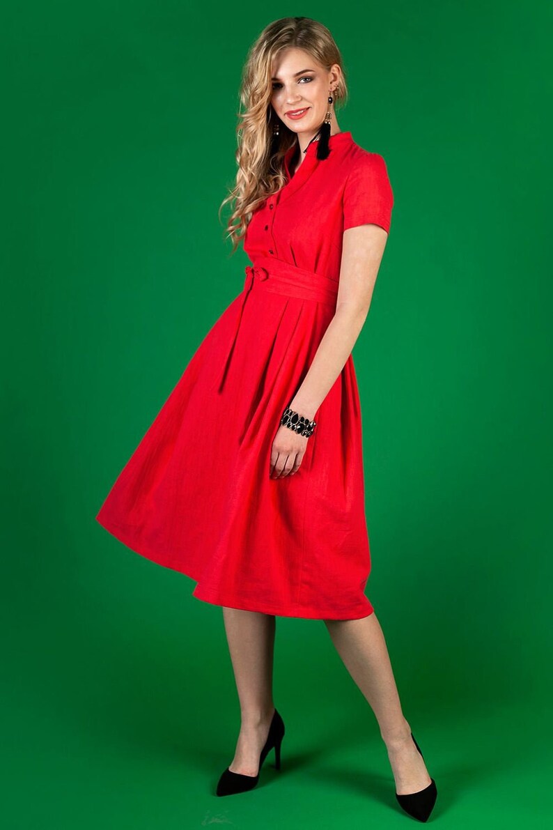 May include A woman wearing a red dress with a belt and short sleeves. She is standing on a green background.