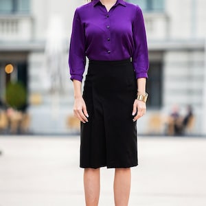 May include: A woman wearing a purple long-sleeved blouse, a black skirt, and burgundy ankle boots. She is standing in front of a building with windows.