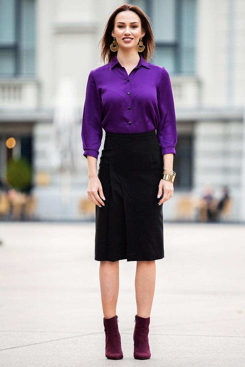 May include: A woman wearing a purple long-sleeved blouse, a black skirt, and burgundy ankle boots. She is standing in front of a building with windows.