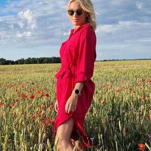 May include: A woman wearing a bright red dress with a belt stands in a field of wheat and red poppies. The dress has long sleeves and a slit. She is wearing sunglasses and a watch. The sky is blue with white clouds.