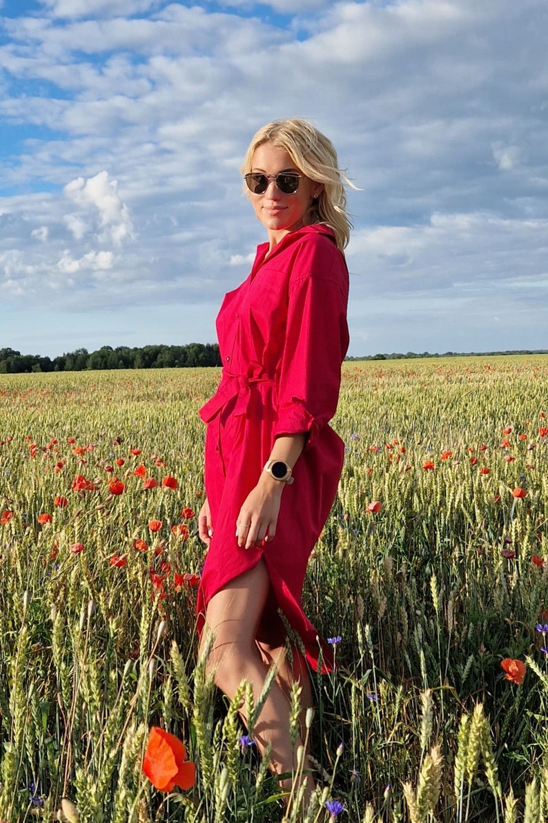 May include: A woman wearing a bright red dress with a belt stands in a field of wheat and red poppies. The dress has long sleeves and a slit. She is wearing sunglasses and a watch. The sky is blue with white clouds.