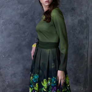 May include: A woman wearing a green dress with a floral print skirt. The dress has a fitted bodice and long sleeves. The skirt is pleated and has a dark green background with colorful flowers.