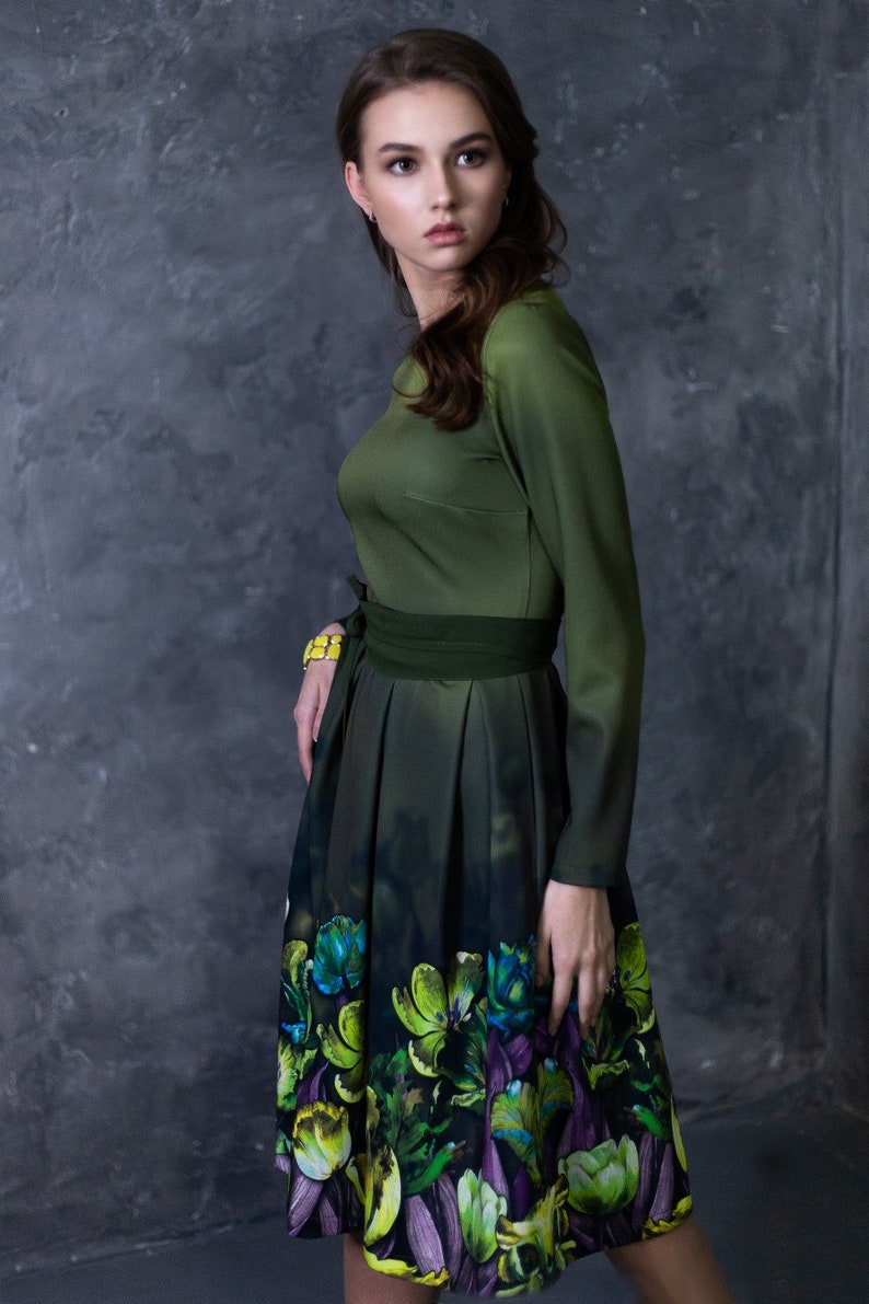 May include: A woman wearing a green dress with a floral print skirt. The dress has a fitted bodice and long sleeves. The skirt is pleated and has a dark green background with colorful flowers.