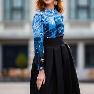 K&ouml;nnte beinhalten: Eine Frau tr&auml;gt eine lang&auml;rmelige Bluse mit blau-wei&szlig;em Muster und einen schwarzen Rock mit hoher Taille. Sie steht vor einem Geb&auml;ude mit gro&szlig;en Fenstern.