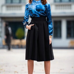 K&ouml;nnte beinhalten: Eine Frau tr&auml;gt eine blau-schwarze gemusterte Bluse mit einem schwarzen, hoch taillierten Rock und schwarzen Stiefeletten.