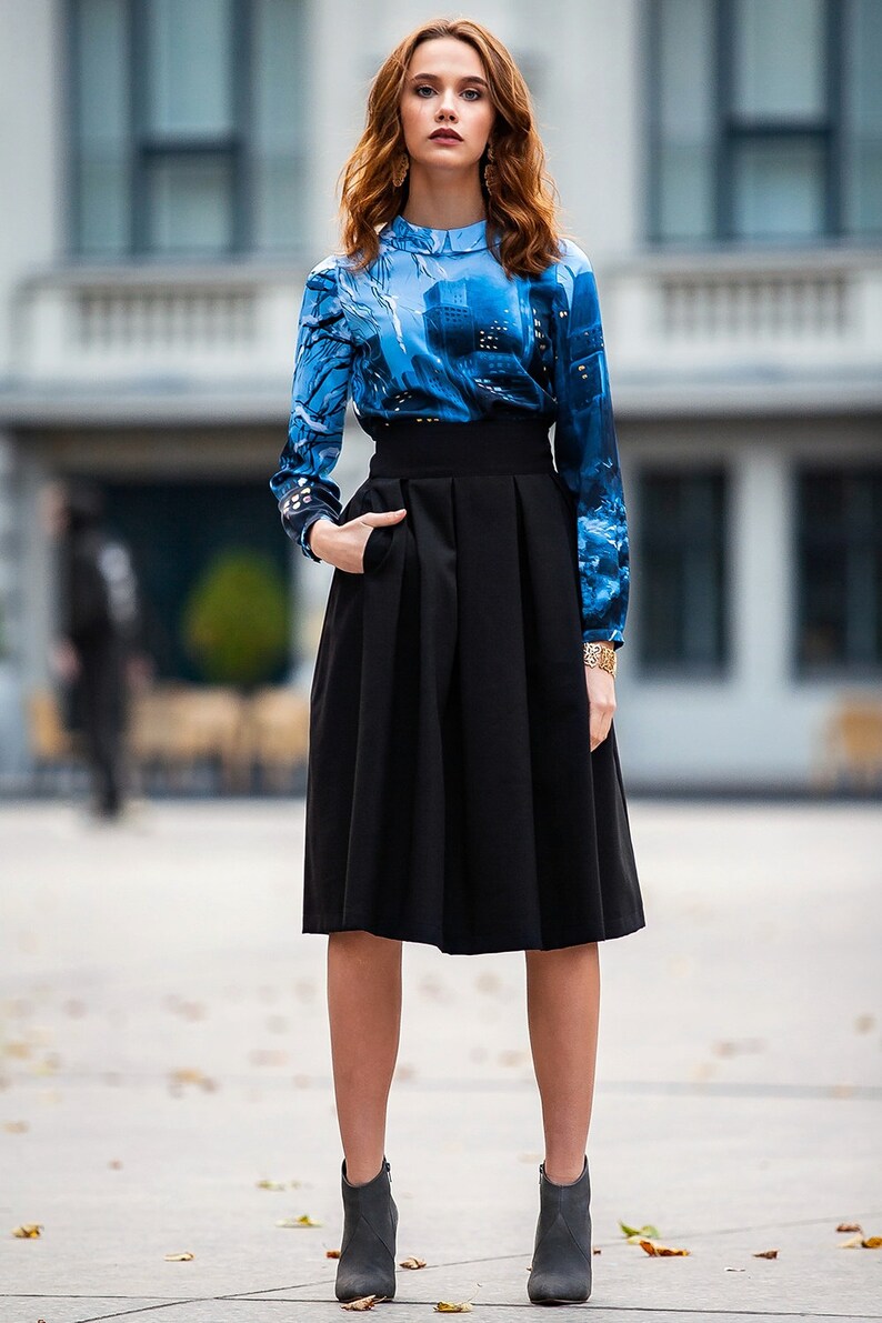 K&ouml;nnte beinhalten: Eine Frau tr&auml;gt eine blau-schwarze gemusterte Bluse mit einem schwarzen, hoch taillierten Rock und schwarzen Stiefeletten.