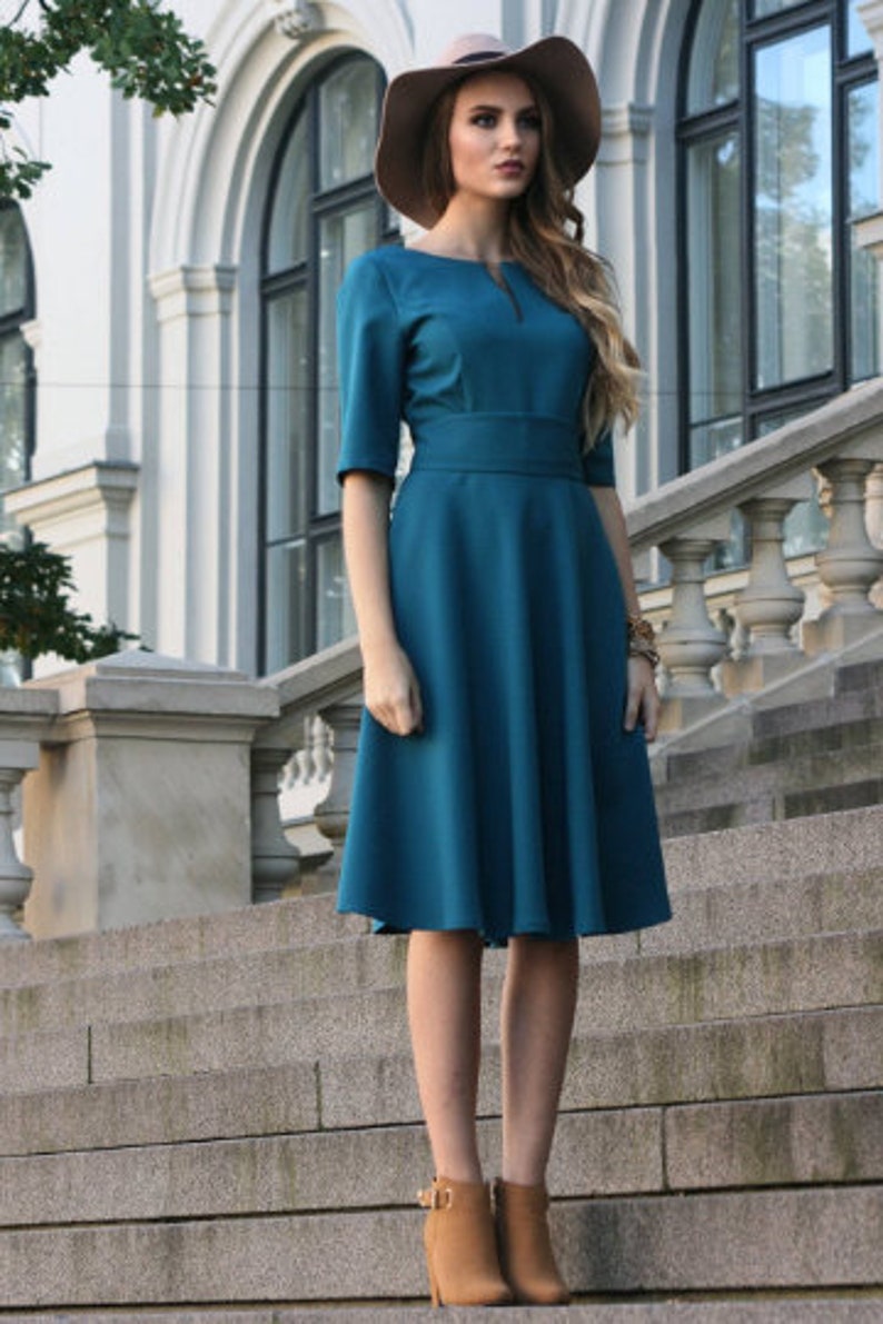 May include: A teal blue dress with a fitted bodice and a flared skirt. The dress has a small keyhole opening at the neckline and a brown belt. The model is wearing brown ankle boots and a brown fedora hat.