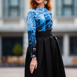 Pu&ograve; includere: Una donna indossa una blusa a maniche lunghe con motivo blu e bianco e una gonna nera. La gonna ha una cintura larga e pieghe.
