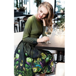 May include: A woman wearing a green dress with a floral print sits at a table with a glass of white wine. The dress has a fitted bodice and a full skirt with a green, purple, and yellow floral pattern.
