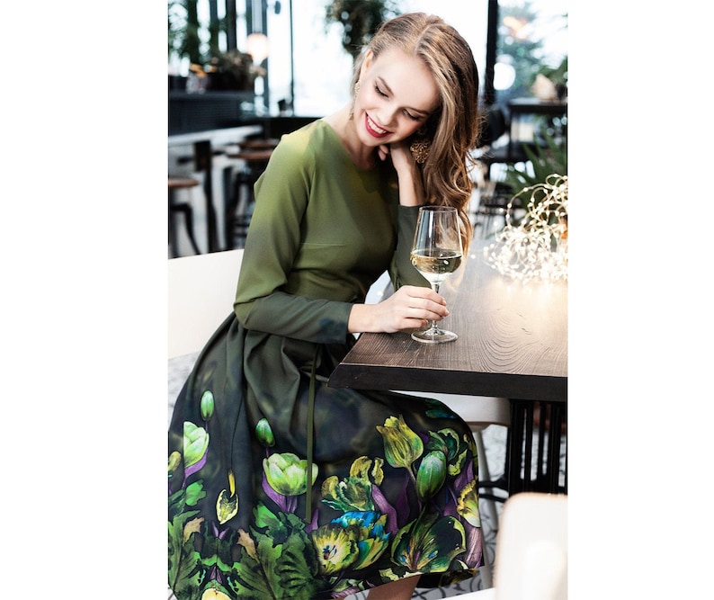 May include: A woman wearing a green dress with a floral print sits at a table with a glass of white wine. The dress has a fitted bodice and a full skirt with a green, purple, and yellow floral pattern.