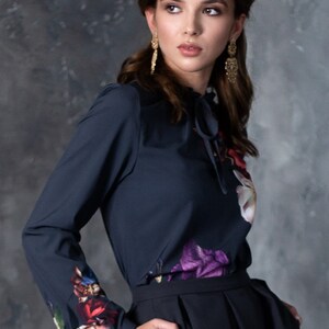 May include: A woman wearing a dark blue long-sleeved blouse with a floral print and a black skirt. The blouse has a tie at the neck and the skirt has pockets.