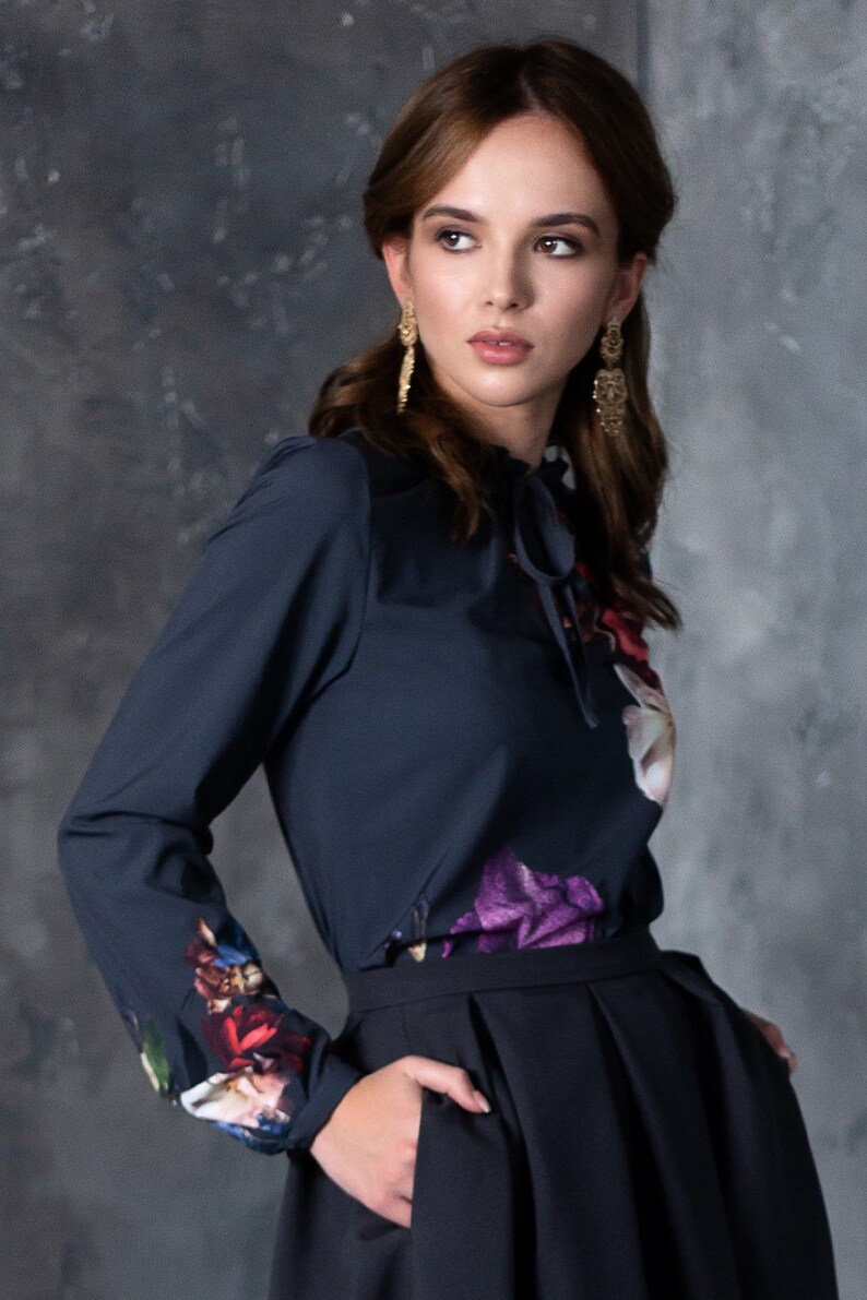 May include: A woman wearing a dark blue long-sleeved blouse with a floral print and a black skirt. The blouse has a tie at the neck and the skirt has pockets.