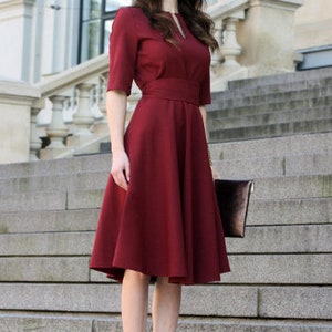 May include: A woman wearing a maroon dress with a fitted bodice and a full skirt. She is standing on a set of stairs and holding a brown clutch.