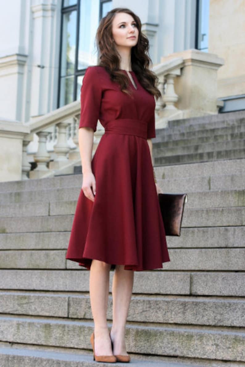 May include: A woman wearing a maroon dress with a fitted bodice and a full skirt. She is standing on a set of stairs and holding a brown clutch.
