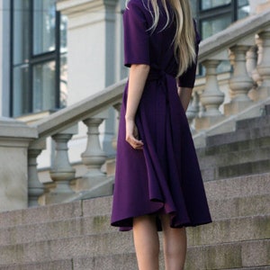May include: A woman wearing a purple midi dress with a tie at the back. She is standing on a set of stone stairs.