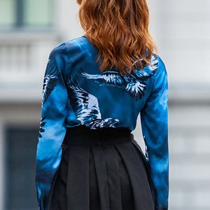 May include A blue silk blouse with a white eagle print. The blouse has long sleeves and a button-up front. The model is wearing a black skirt.