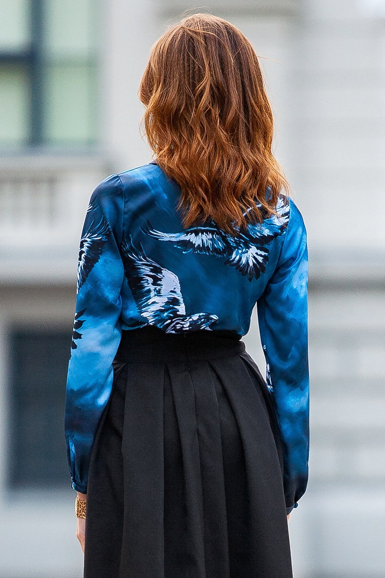 May include A blue silk blouse with a white eagle print. The blouse has long sleeves and a button-up front. The model is wearing a black skirt.
