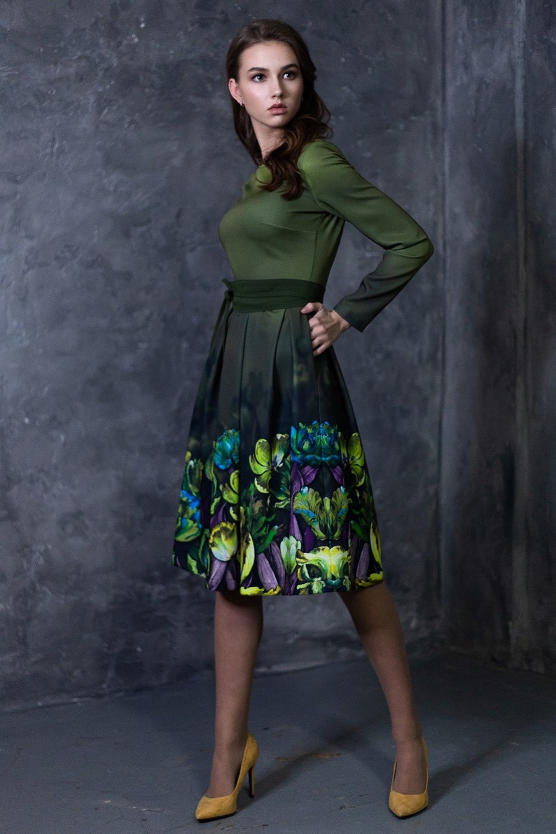 May include: A woman wearing a green dress with a floral print skirt and yellow heels. The dress has a green bodice and a dark green belt.