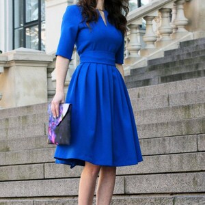 May include: A woman wearing a royal blue knee-length dress with a pleated skirt and a v-neck. She is holding a floral clutch and wearing black heels.