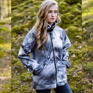 Puede incluir: Una chaqueta gris y blanca con estampado, cierre de cremallera y cuello alto. La chaqueta la lleva puesta una persona de pie en un entorno boscoso.