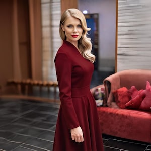 Könnte beinhalten: Eine Frau in einem weinroten Kleid mit langen Ärmeln posiert in Innenräumen. Das Kleid hat ein eng anliegendes Oberteil und einen ausgestellten Rock. Der Hintergrund umfasst ein rotes Sofa mit roten Kissen und einen dunklen Fliesenboden.