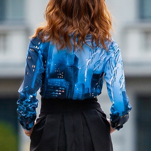 K&ouml;nnte beinhalten: Eine Frau tr&auml;gt eine blau-wei&szlig;e gemusterte Bluse mit einem schwarzen Rock. Die Bluse hat ein Stadtbild-Design.