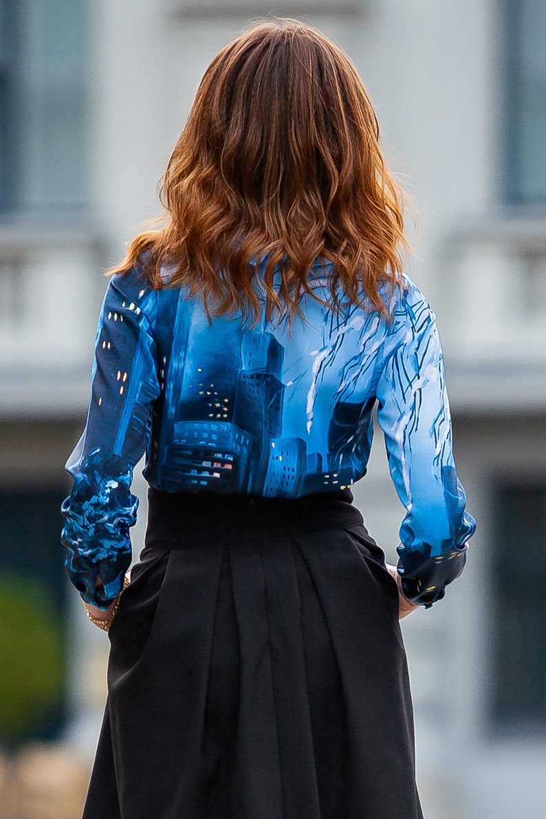 K&ouml;nnte beinhalten: Eine Frau tr&auml;gt eine blau-wei&szlig;e gemusterte Bluse mit einem schwarzen Rock. Die Bluse hat ein Stadtbild-Design.