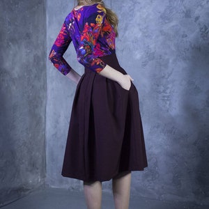 Pu&ograve; includere: Una donna indossa una blusa con stampa floreale viola e rossa, una gonna midi bordeaux e stivaletti neri con tacco.