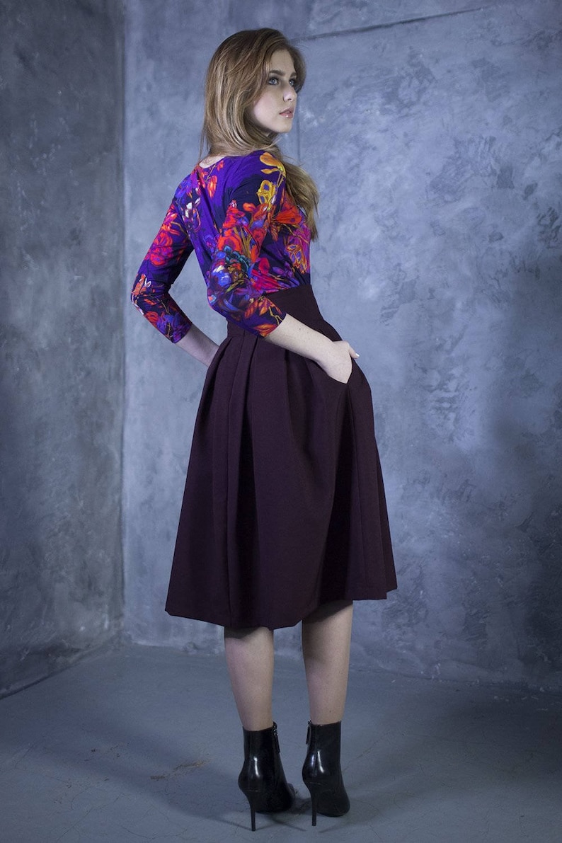 Pu&ograve; includere: Una donna indossa una blusa con stampa floreale viola e rossa, una gonna midi bordeaux e stivaletti neri con tacco.